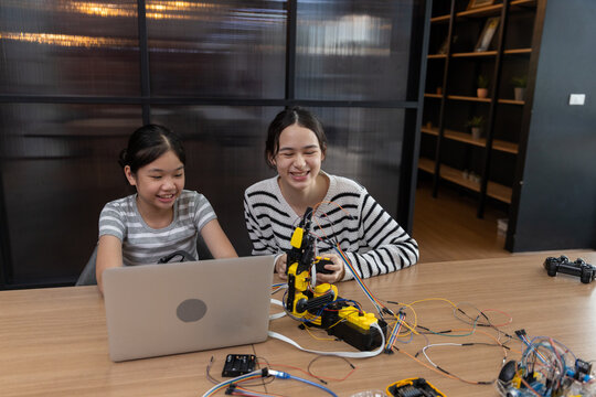 Happy young Asian female student building yellow robot arm using laptop computer on wooden table during fun education lesson