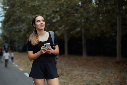 Captivating Woman Explorer Standing On Pathway Holding Her Smartphone Outdoor. Smiling woman looks up while standing on a forest road holding her mobile phone