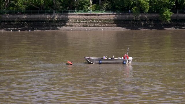 Single empty motorboat gently rocks on a muddy brown river near a stone embankment lined with trees on a calm day.