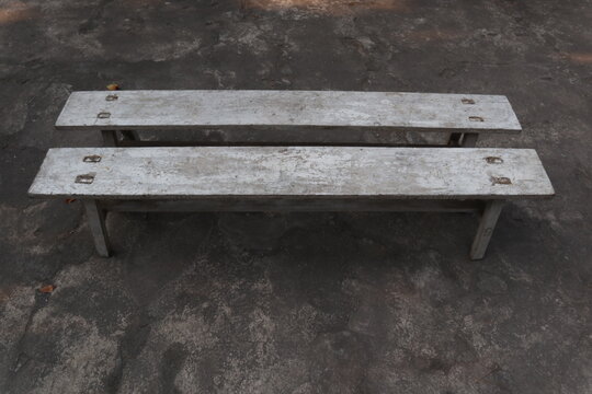 Two long, weathered wooden benches with faded white paint placed parallel on a concrete yard in a rural village in Java, Indonesia.