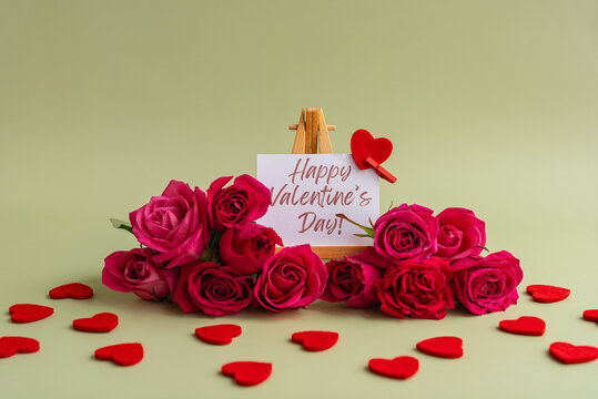 Happy Valentine's Day greeting card displayed on wooden easel surrounded by vibrant pink roses and decorative red heart shapes on soft pastel background. Romantic floral arrangement with elegant