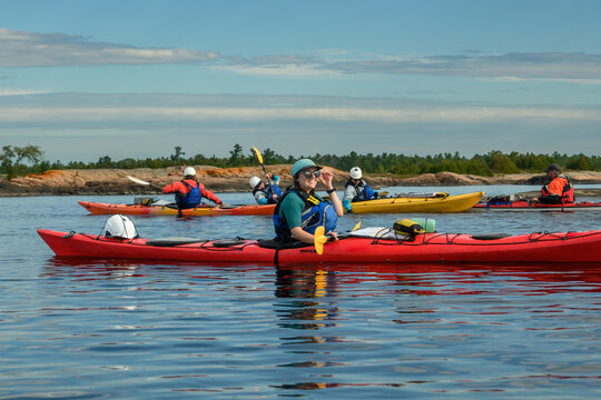 Smiling woman adjusting her sunglasses while sitting in a long red sea kayak with paddling group behind, blue sky copy space, Georgian Bay, Ontario