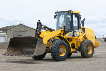 Yellow front loader with a large bucket on an outdoor construction site, surrounded by gravel and industrial buildings. © Patagonia pics