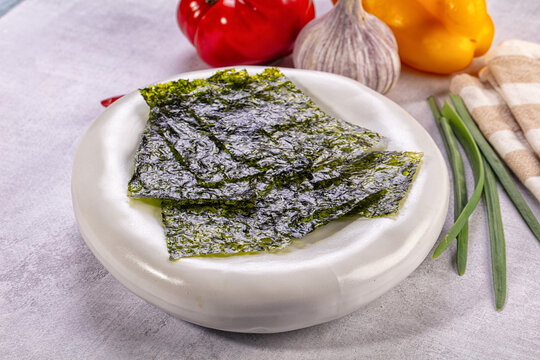Crispy green nori sheets on white ceramic plate with red bell pepper, yellow bell pepper, garlic bulb, and green onions in background