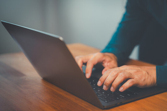 Close up view adult man using laptop computer keyboard while working at wooden office desk using internet technology for digital communication and work.