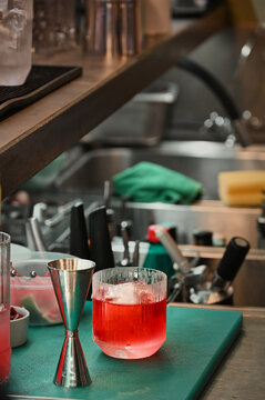 Close-up of a low glass filled with a red cocktail with ice and a metal jigger on a green cutting board in a professional bar workspace, bartending and mixology concept.