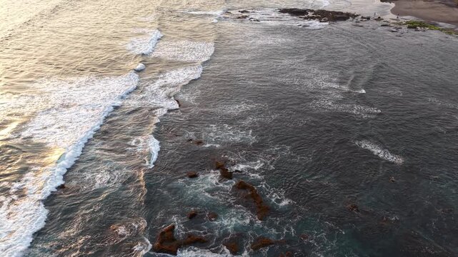 Golden sunset light illuminates an aerial view of Kelegung Beach shoreline with crashing waves breaking over rocky reefs, creating textured surf, coastal patterns and a tranquil tropical seascape mood