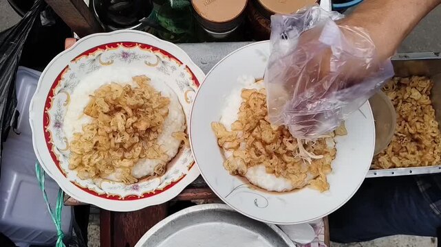 Top-down view of a street food vendor preparing Indonesian Bubur Ayam (Chicken Porridge) by adding shredded chicken and green onions