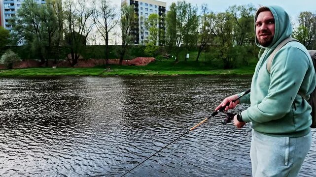 Young fisherman in a hoodie using a spinning rod and reel on a city river during daytime