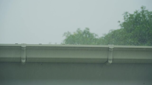 Heavy rain falling on house roof with green trees background during tropical rainy season.