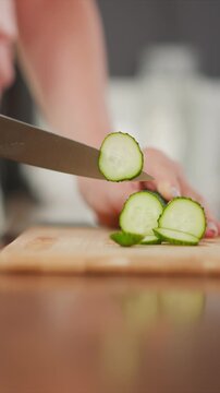 Woman slices cucumber into thin rounds on wooden cutting board. fresh green vegetable is cut with precision revealing its interior texture and seeds. Soft lighting highlights smooth surface