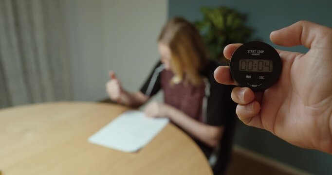 Woman writes a test while a hand in the foreground measures the time with a stopwatch.