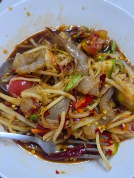A close-up of spicy Thai som tam with fresh raw shrimp, shredded green papaya, tomatoes, chilies, and a glossy tangy dressing on a white plate.