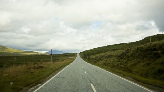 First person dashcam view shows an asphalt road crossing rolling green hills and open moorland. Cloudy skies and distant hills create a scenic countryside drive in Derbyshire England.
