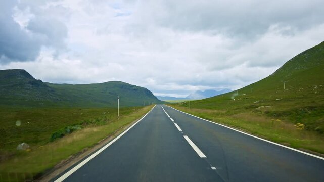 First person dashcam view shows driving along a smooth asphalt road through rolling green hills and open moorland. Cloudy skies create a scenic countryside landscape in Derbyshire England.