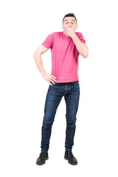 Full body of shocked male covering mouth with hand and looking at camera while standing with hand on waist