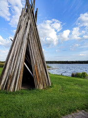 Wooden teepee structure standing on green grass field by the lake against blue sky with white clouds © Lidya