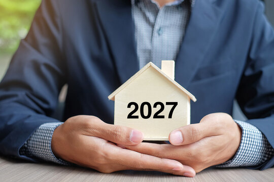 Businessman hands holding wooden House model with 2027 New Year text. Property insurance and real estate concepts
