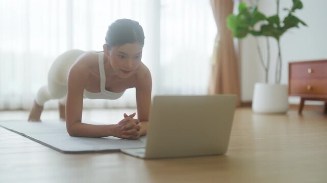 Young Asian woman in sportswear doing plank on training mat while watching online workout tutorials on laptop in the living room. Sport and recreation concept