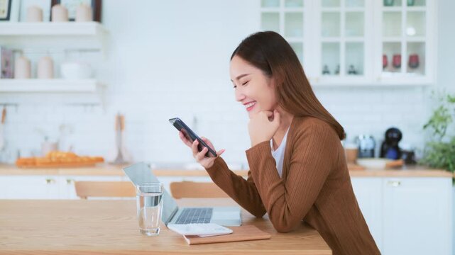 Happy young asian woman relax in domestic kitchen at home, using smartphone, smiling girl use cell phone chatting, browse wireless internet on gadget, shopping online from home