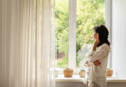 Mature woman in a white blouse stands by large, light-filled window with sheer curtains, gazing at green trees. She feels at peace, experiencing quiet solitude. Authentic slow living