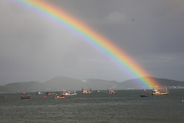Obraz premium A vibrant rainbow arching over the sea with local fishing boats against a dramatic stormy sky in Sattahip, Chonburi, Thailand.