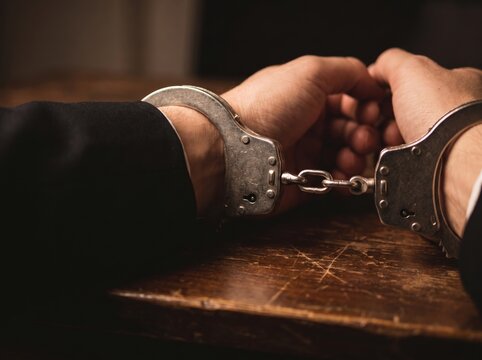 Close up of a businessman in a suit with handcuffed hands sitting at a wooden table after being arrested