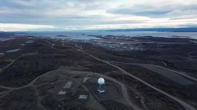 Continuation of aerial drone pull back shot traveling backward high above radar dome revealing expansive tundra bedrock and Frobisher Bay waters