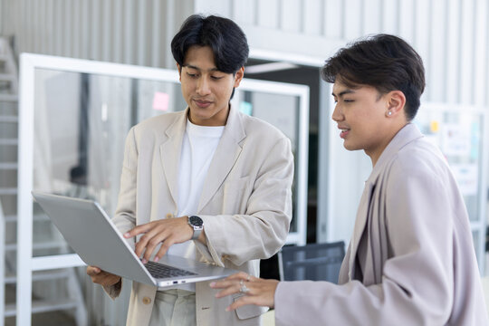 Two confident Asian professional men working together with laptop computer in modern bright corporate office business meeting