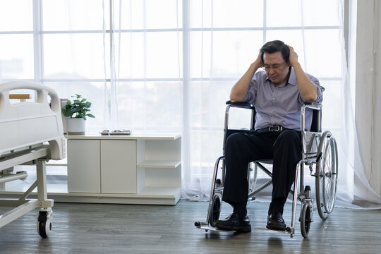 Depressed senior man sitting in wheelchair holding head with sad emotional expression feeling lonely inside hospital room near bed and window