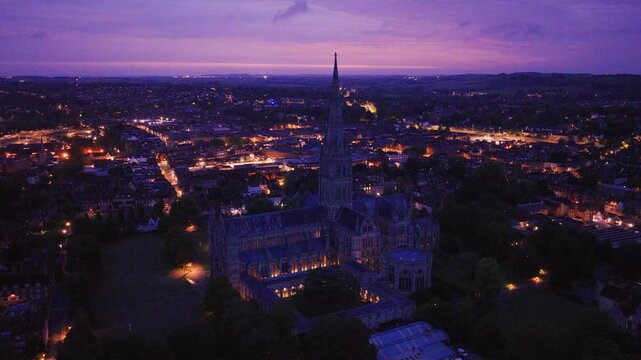 Aerial view of Salisbury Cathedral at blue hour with its illuminated Gothic architecture rising above the city lights and surrounding Wiltshire landscape