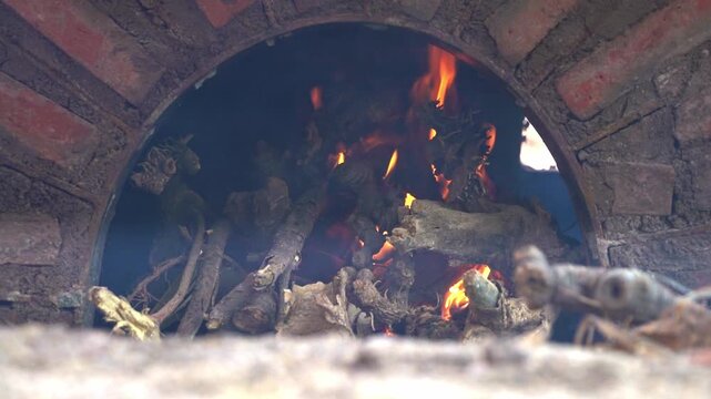Close up shot of stacked firewood slowly catching fire inside a dark, traditional roasting pit, with small flames and smoke beginning to build as the oven heats up