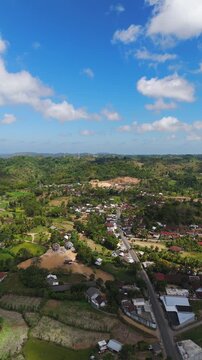 Sade village Lombok Indonesia aerial drone pull bac, lush green hills tropical countryside rooftops and winding road vertical shot.