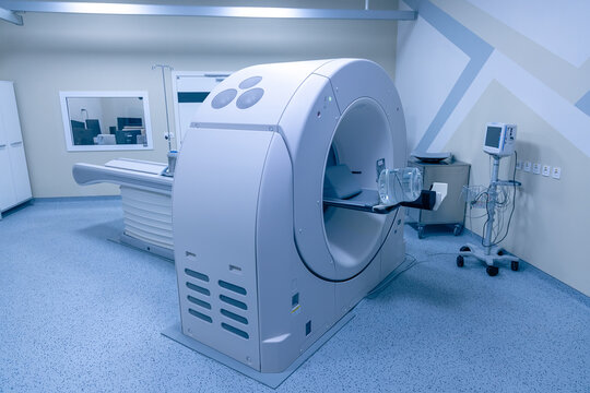 A computed tomography scanner with a patient table stands in a new diagnostic room. A monitor on a stand waits by the wall, and a control room is visible through the window. The scanner is ready