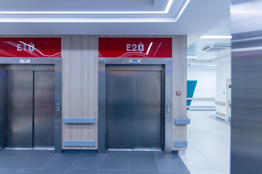 Two elevators with steel doors stand side by side in the lobby of a new hospital. Red signs with numbers mark each lift above the doors. A bright corridor leads away to the right