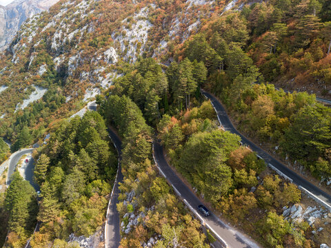 Lovcen Serpentine Montenegro switchback road snakes through dense forest and rocky mountain terrain. Winding asphalt bends around cliffs and autumn foliage, with one car moving along scenic route.