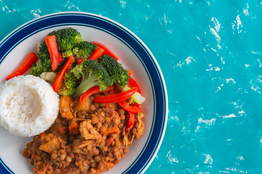 Vibrant meal sits on bold blue plate. White rice nestled beside lentils and veggies. Red peppers, broccoli, and chicken add color. Bright turquoise background enhances freshness