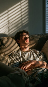 Man Resting on Sofa in Sunbeams: A young man is peacefully resting on a comfortable sofa, illuminated by the warm, dramatic streaks of sunlight filtering through window blinds.