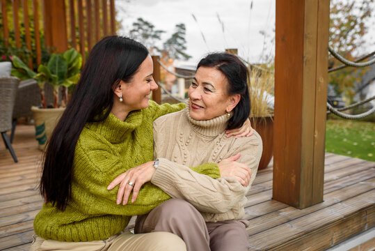 Mature daughter with older mom sitting on wooden patio, hugging. Mother's day concept.