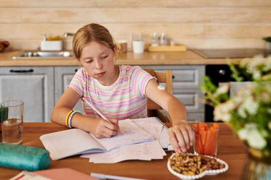 Caucasian kid girl with continuous glucose monitor writing in notebook doing homework and eating healthy snacks. Perfect for diabetes management and pediatric healthcare campaigns