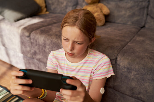 Caucasian kid girl with diabetes wearing glucose monitor sensor on arm playing handheld video game console at home. Suitable for juvenile diabetes management advertising and health campaign