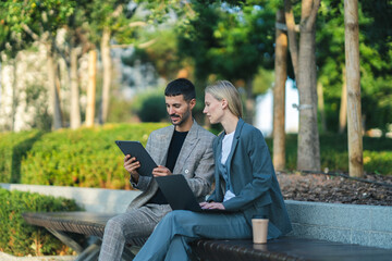 Fototapeta premium Two business professionals discussing work on a tablet and laptop in a park setting