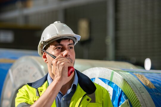Technician controlling metal sheet machine performance operation system control setting in manufactory Portrait of Hispanic male engineer talking radio transceiver near steel coils at warehouse