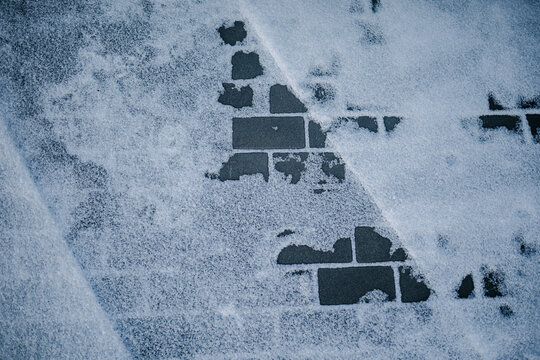 Snow rests lightly on a paved sidewalk surface. Square tiles form a regular, grid-like pattern.