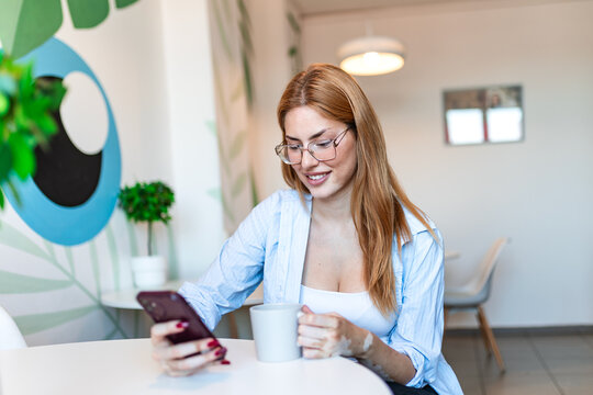 Coffee break at work. Mobile communication and rest time, Business lifestyle and technology concept. Young smiling businesswoman sitting at a cafe table, holding phone and cup.