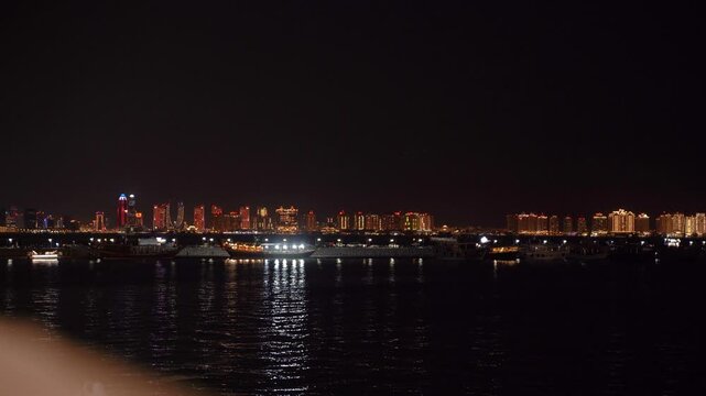 Traditional qatari dhow boats sailing along the waterfront with the illuminated modern doha city skyline reflecting on the calm waters of the persian gulf during a clear night in qatar