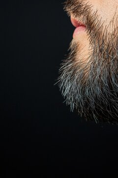 Close-up of a man's lower face showing a thick, dark beard with some gray hairs and pink lips against a black background.