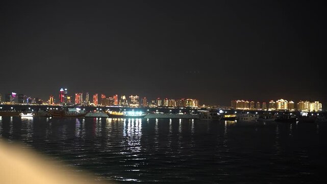 Qatar's capital city, doha, shimmering at night with its modern skyscrapers and vibrant lights reflecting on the calm sea, while traditional dhow boats and yachts are docked in the bay
