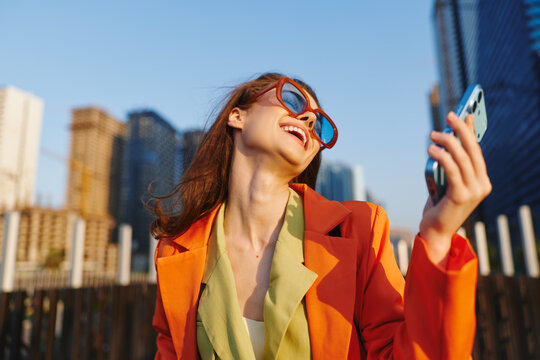 Happy woman in orange jacket and sunglasses laughing while looking up, holding smartphone against city skyline. Concept of euphoria, joy, and living in the moment with mobile technology.