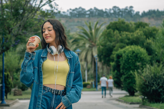 A relaxed young woman listening to music and drinking a takeaway beverage while walking down the street